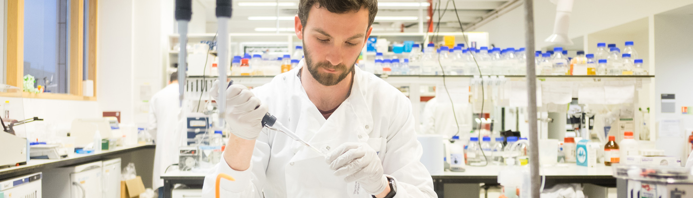 Researcher wearing whtie lab coat surrounded by blue capped bottles squeezing pipette into tube